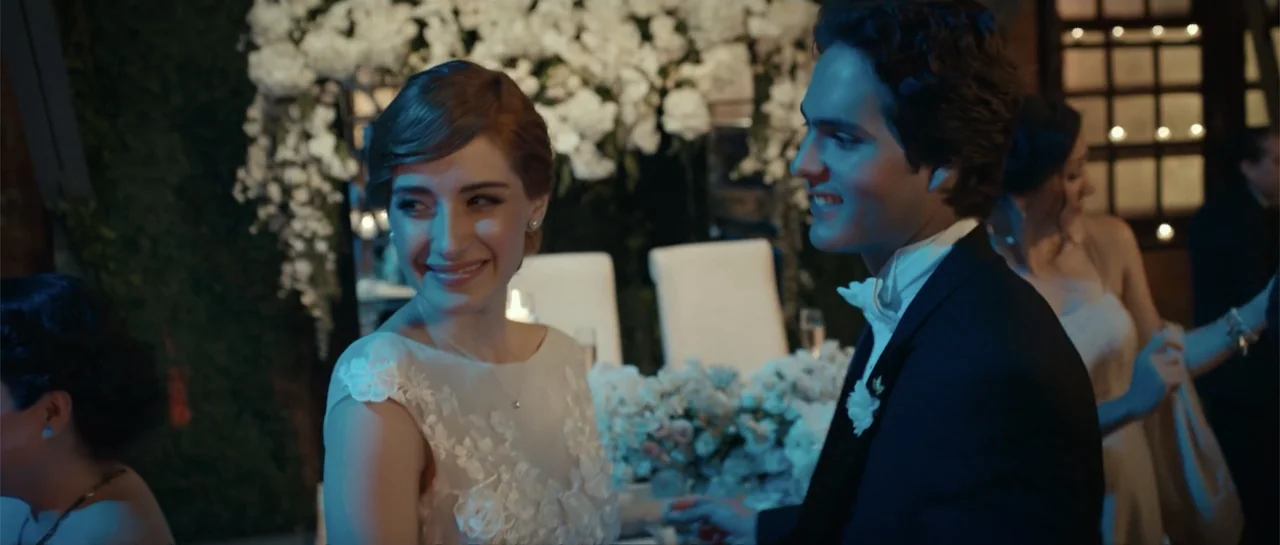 Cindy La Regia (2020) movie still: medium — A dimly lit event space filled with white floral arrangements and warm string lighting. A bride and…