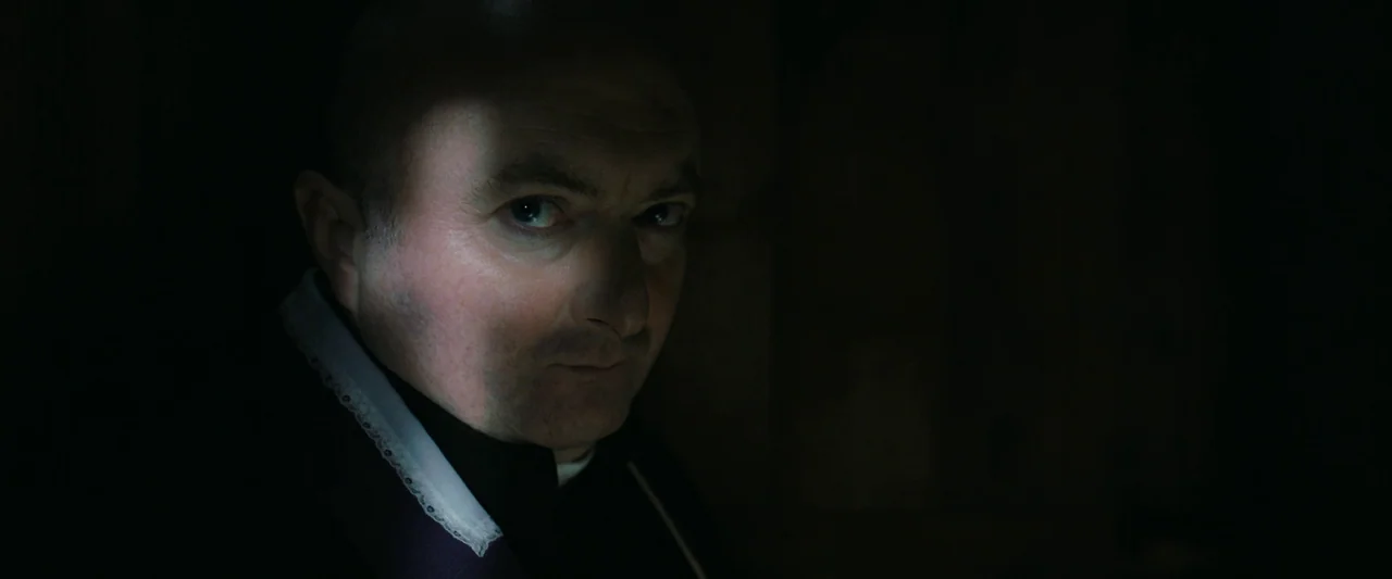 The Banshees of Inisherin (2022) movie still: closeup — A dark confessional booth with the subject positioned in the frame's light source. The man…