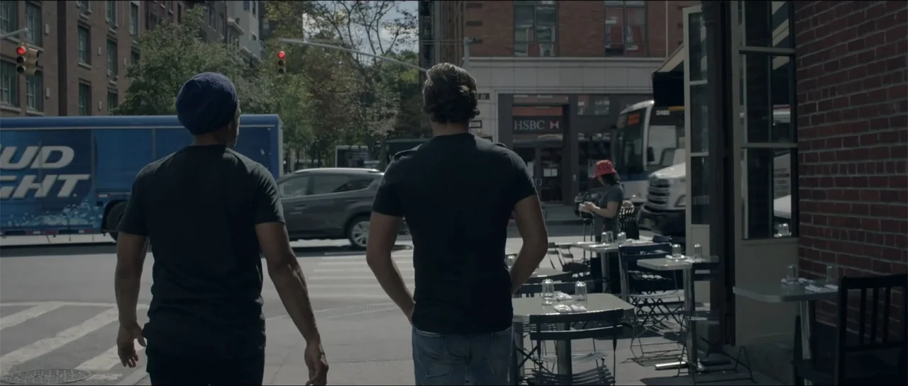 All She Wrote (2018) movie still: fullbody — A city sidewalk with outdoor dining tables sits adjacent to a busy street intersection. Two men walk…