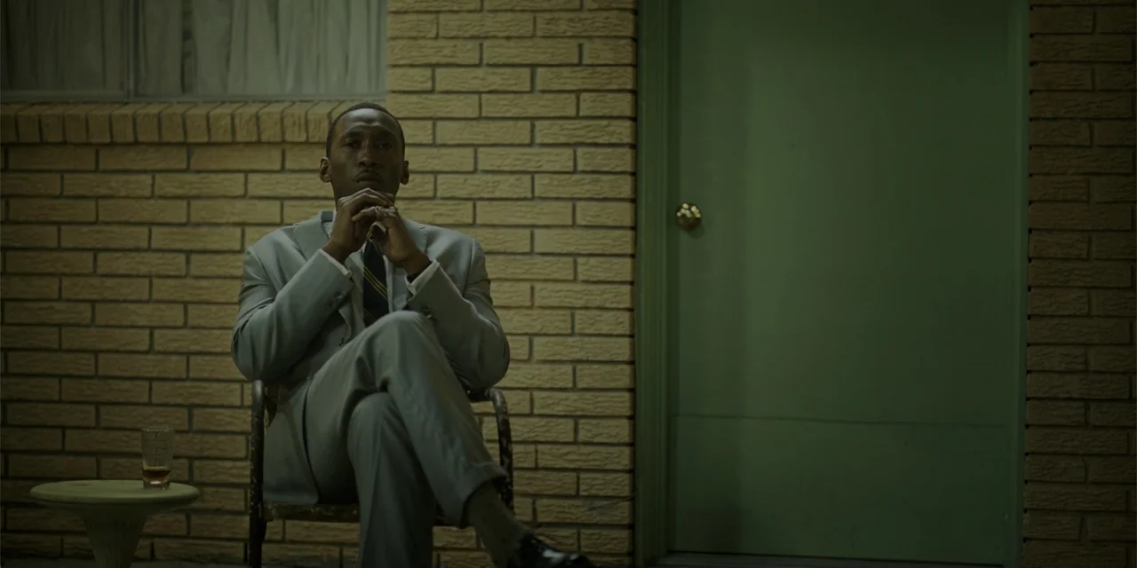 Green Book (2018) movie still: medium — A beige brick exterior wall sits next to a closed green door. A man in a grey suit sits in a metal chair.