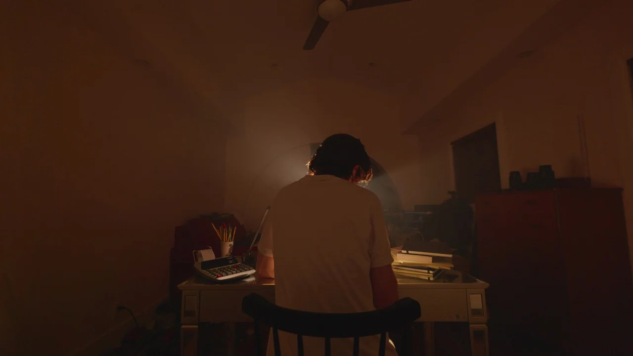 Bo Burnham: Inside (2021) movie still: medium — A dark, dimly lit space features a person working at a desk. The subject sits facing away from the…