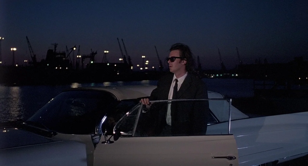Ariel (1988) movie still: medium — A man stands next to a white vintage convertible at night near a harbor. Several construction cranes and…