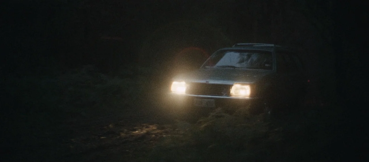 Force of Nature: The Dry 2 (2024) movie still: medium — A station wagon sits on a dark, dirt track surrounded by dense dark vegetation. The car's…