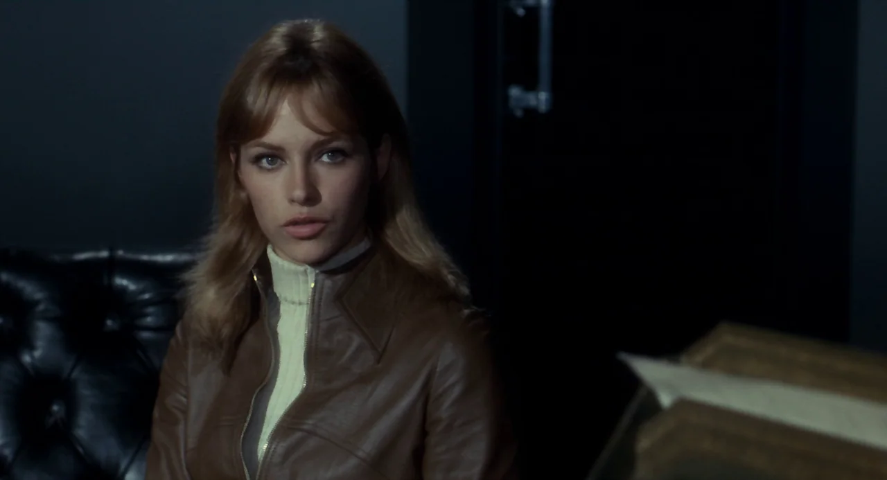 Le Samouraï (1967) movie still: medium — A dark office environment with black walls and a tufted leather chair. A young woman with blonde hair sits…