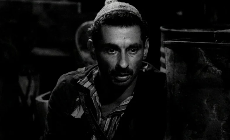 Cairo Station (1958) movie still: medium — A dark, cramped space filled with large storage containers in deep shadow. A man with a mustache looks…