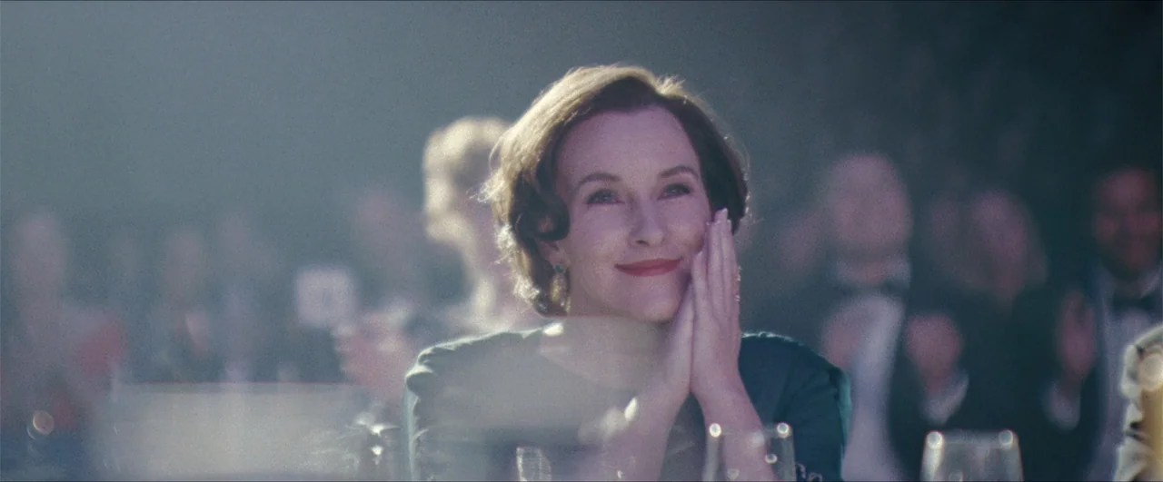 Better Man (2024) movie still: medium — A crowded banquet hall filled with blurred guests seated at round tables. A woman in a green dress sits at a…