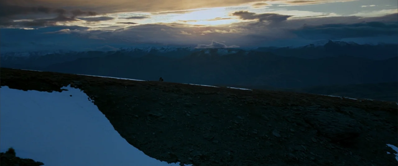 10,000 BC (2008) movie still: establishing — A vast mountain range stretches under a heavily clouded sunset sky. Sparse snow patches cover the rocky…