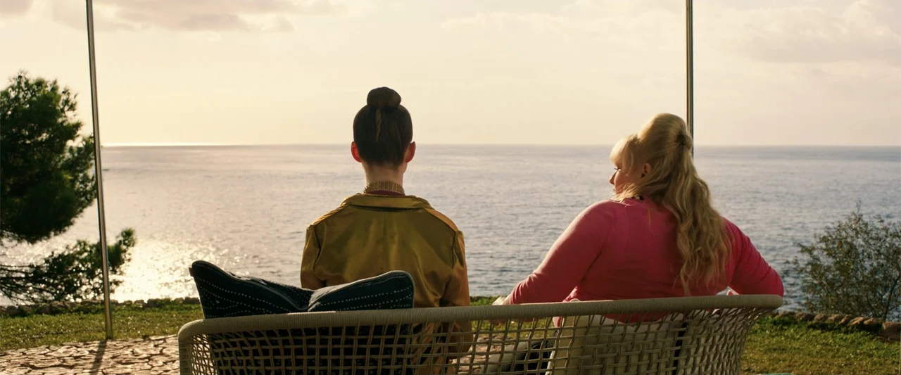 The Hustle (2019) movie still: medium — A sunny patio overlooks the calm ocean during the golden hour. Two women sit on a wicker bench looking at…