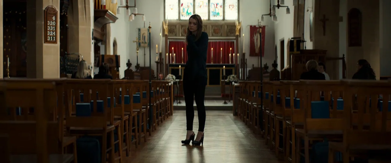 Silver Haze (2023) movie still: fullbody — A traditional church interior with wooden pews and a stained glass window backdrop. A young woman stands…