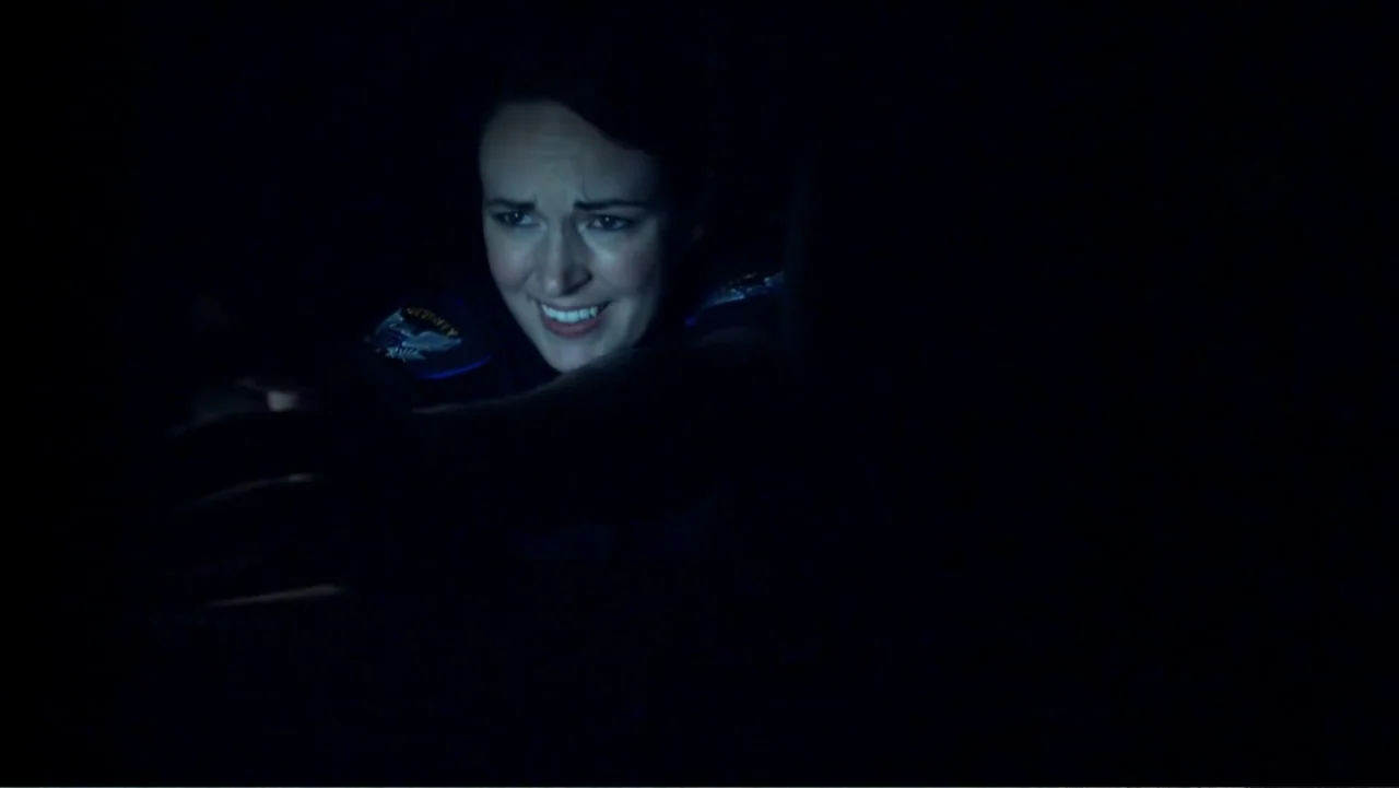 Abandoned Dead (2015) movie still: closeup — A dimly lit interior space with the subject appearing against a pitch black background. A woman in a…