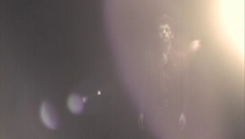 The Tracey Fragments (2007) movie still: medium — A person stands in a dimly lit, nondescript area with heavy video scanline artifacts. Strong light…