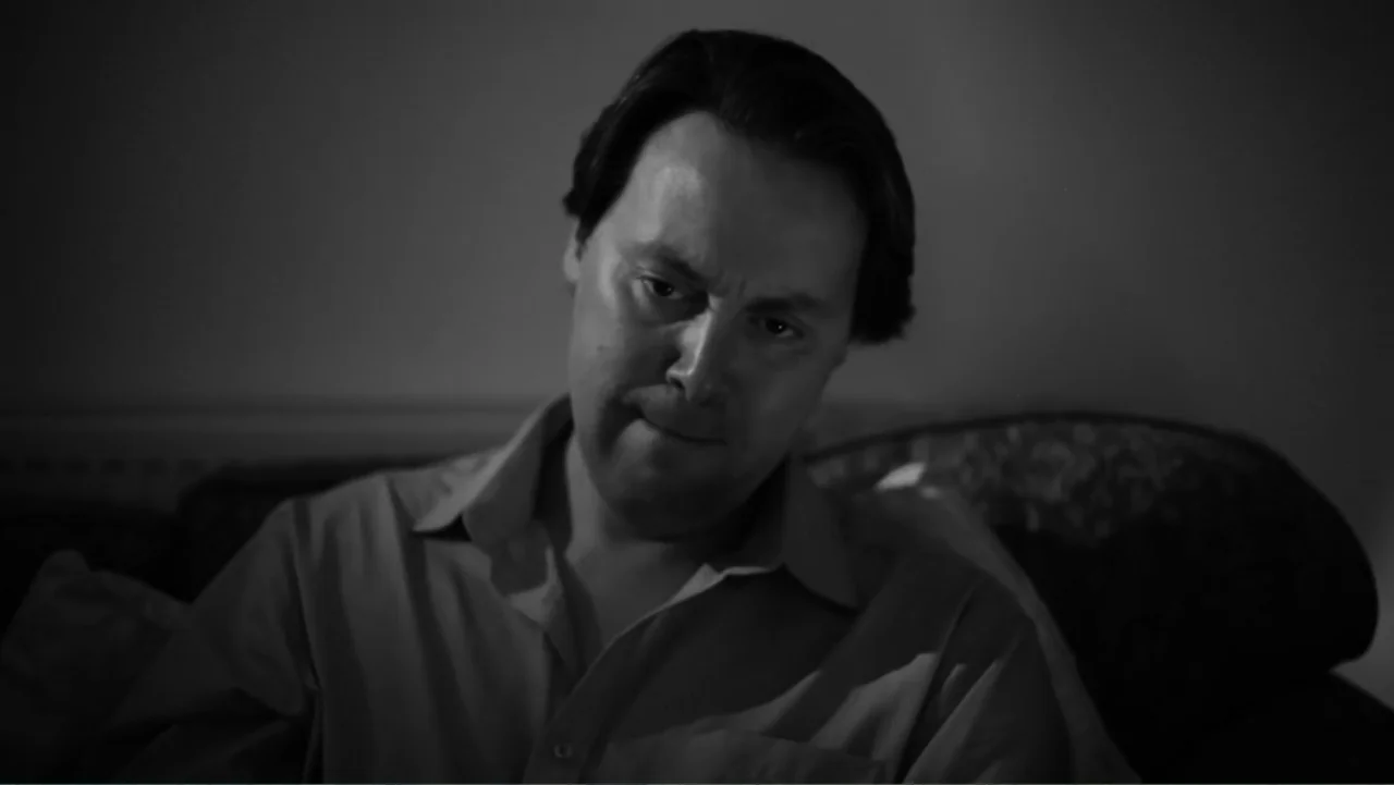 Creditors (2025) movie still: medium — A dimly lit domestic interior features a man sitting on a patterned sofa. The man is captured from the torso…