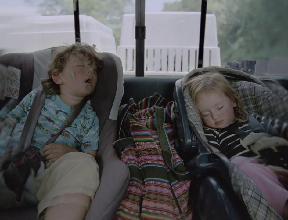 Post Tenebras Lux (2012) movie still: medium — Two children sleep in car seats positioned in the back row of a vehicle. A woven blanket is tucked…