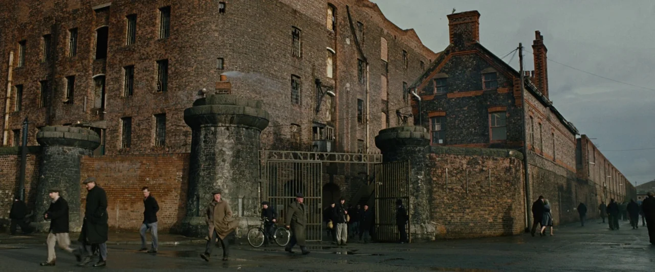 Across the Universe (2007) movie still: wide — A massive industrial brick building with circular stone towers stands behind a large metal gate.…