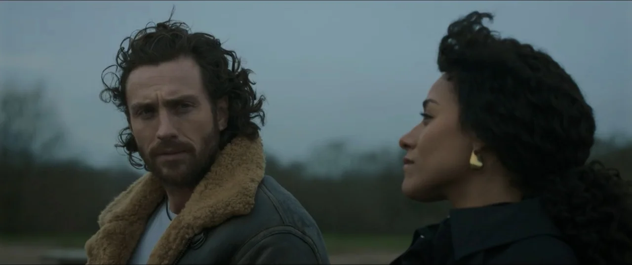 Kraven the Hunter (2024) movie still: medium — Two individuals stand against a blurry, overcast outdoor backdrop with muted natural colors. The man…