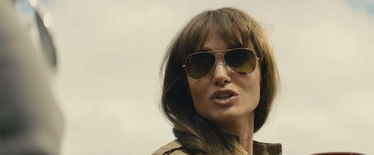 Those Who Wish Me Dead (2021) movie still: closeup — An outdoor location with an indistinct bright sky backdrop. A woman in a tan jacket is in a…