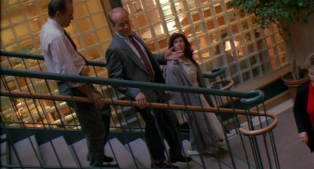Raising Cain movie still: medium — A stairwell features glass brick walls and a metal handrail system. Three people descend the stairs while the…