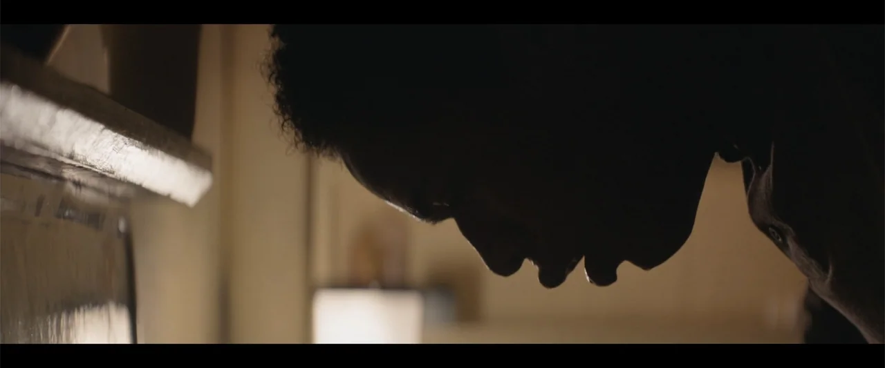 It Comes at Night (2017) movie still: closeup — An interior space contains a person partially illuminated from a light in the background. The person…
