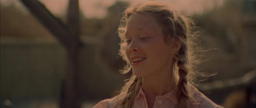 3 Women (1977) movie still: closeup — A rural setting with a soft-focus background of wooden structures and light. A teenage girl with braided hair…