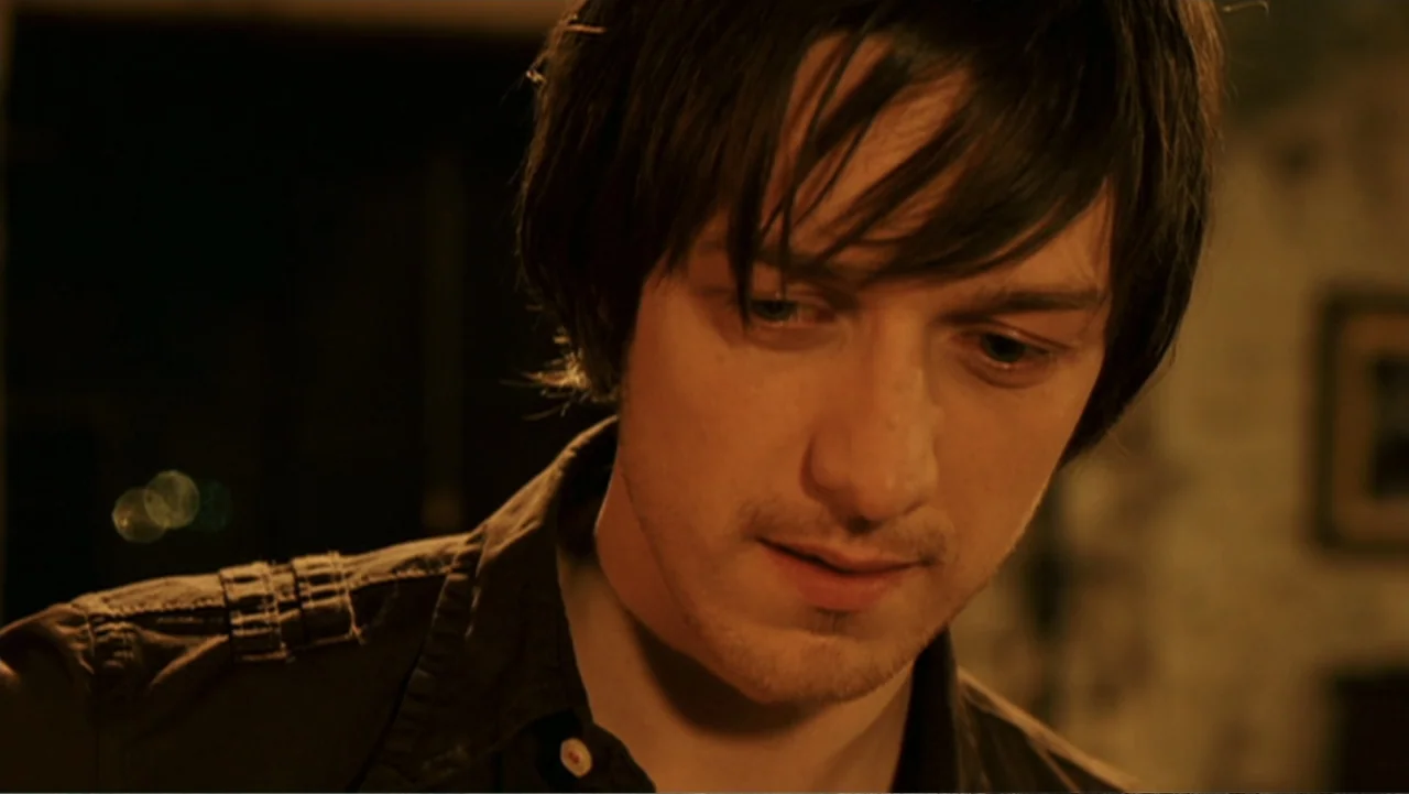 Penelope (2007) movie still: closeup — A dimly lit indoor space with blurred background details. A young man with dark hair looks down with a…