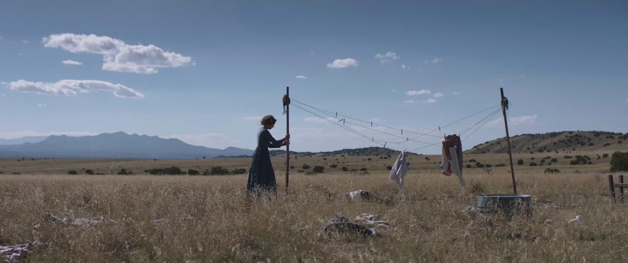 The Wind (2018) movie still: wide — A woman stands alone in a wide, dry, grassy plain beneath a clear blue sky. She adjusts a clothesline pole while…