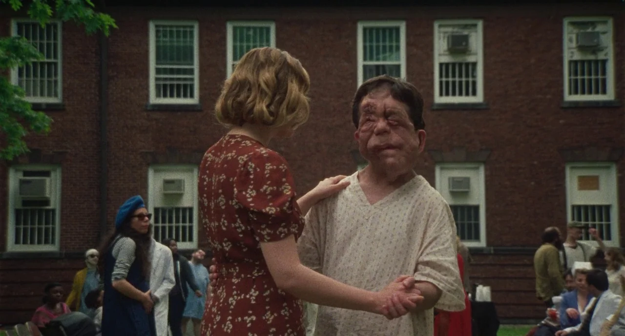 Chained For Life (2018) movie still: medium — An outdoor courtyard with a large red brick building in the background. A woman in a floral dress…