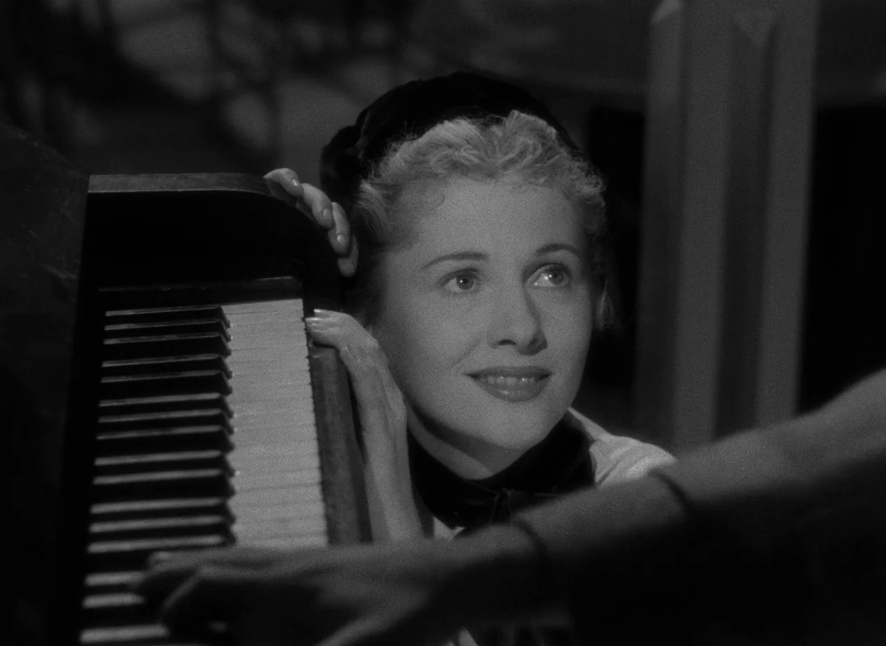 Letter From An Unknown Woman (1948) movie still: closeup — A dimly lit room focuses on a woman resting her head against a piano. Her face is…