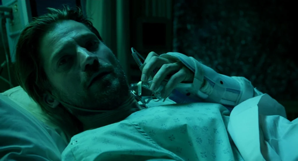 Mama (2013) movie still: closeup — A dark hospital room at night illuminated by cool green light. A man with an injured arm lies in bed holding an…