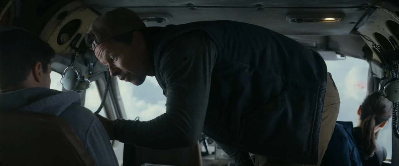 Flight Risk (2025) movie still: medium — An interior cockpit features worn control panels and headphones hanging in the workspace. A man leans over…