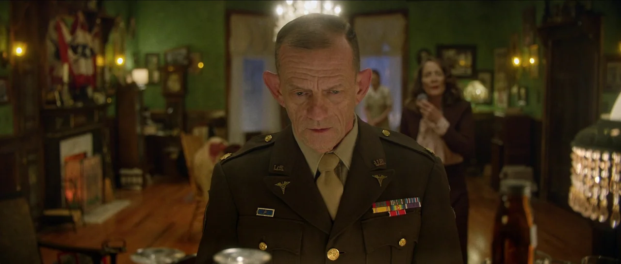 Brooklyn 45 (2023) movie still: medium — A dark green parlor interior filled with wood furniture and decor from the mid-century. An elderly man in…