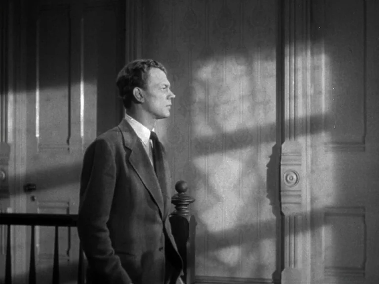 Shadow of a Doubt (1943) movie still: medium — A man stands in a hallway or landing with patterned wallpaper and dark wood trim. Hard sunlight cuts…
