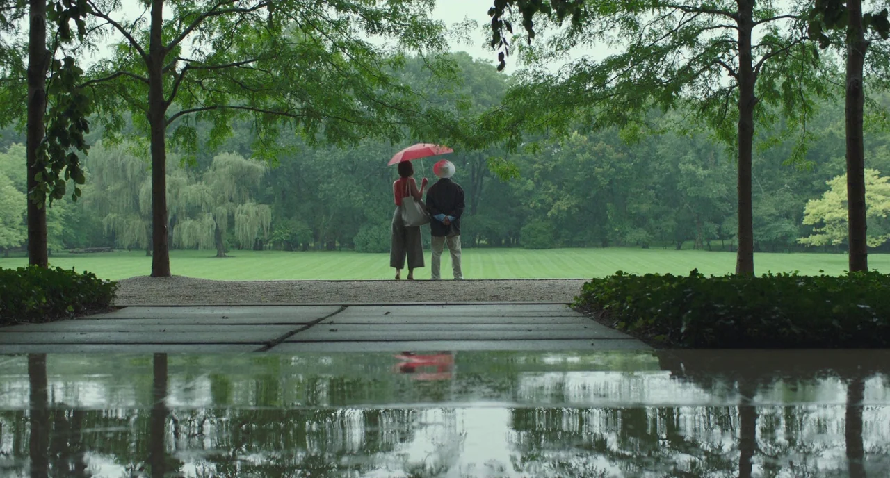 Columbus (2017) movie still: wide — A wet concrete walkway leads to an expansive manicured lawn framed by tall trees. Two people stand under a…