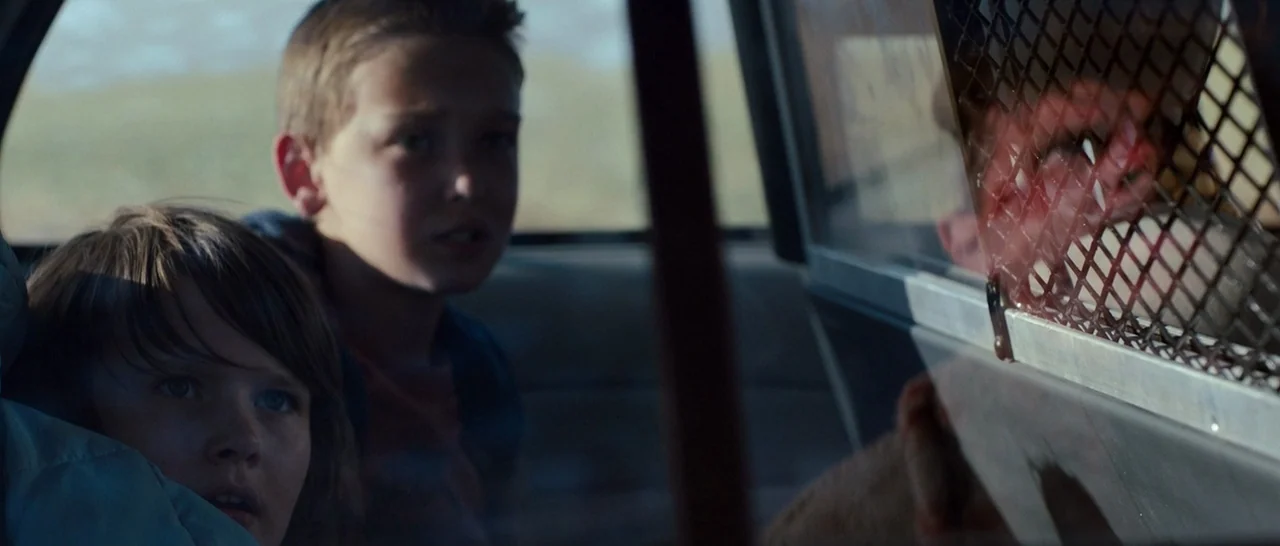 Cop Car (2015) movie still: medium — The interior of a police cruiser features a metal security grate separating the seats. Two children sit in the…