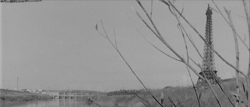 I Vampiri (1957) movie still: wide — A rural riverbank features dry grass and thin bare branches in the foreground. The Eiffel Tower rises…