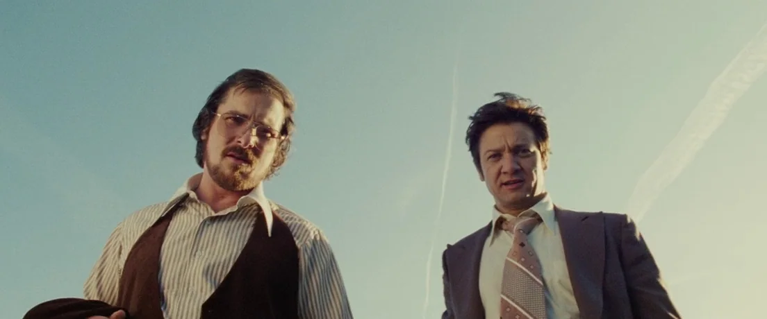 American Hustle (2013) movie still: medium — Two men stand against a clear blue sky in an open, sunlit outdoor area. They look directly down at the…
