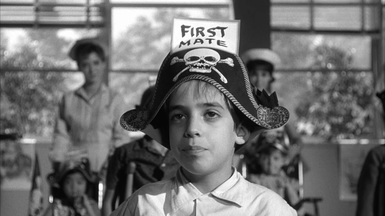 The Naked Kiss (1964) movie still: closeup — A young boy sits in a classroom filled with children wearing pirate hats. The boy stares directly ahead…