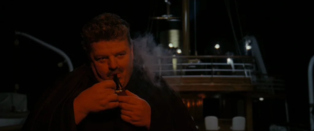 The Brothers Bloom (2008) movie still: medium — A dark deck of a ship at night with metal railings and masts. A heavy-set man lights a pipe while…