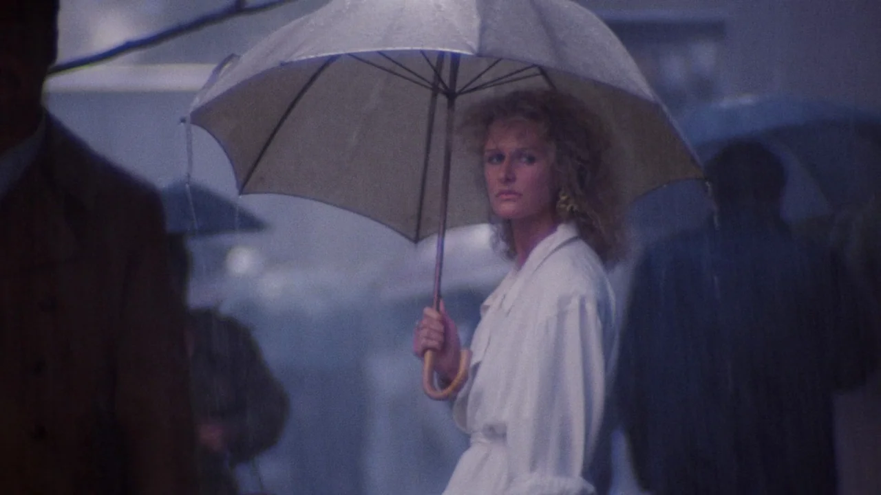 Fatal Attraction (1987) movie still: medium — A city street at night during a rainstorm with blurred pedestrian figures nearby. A blonde woman…