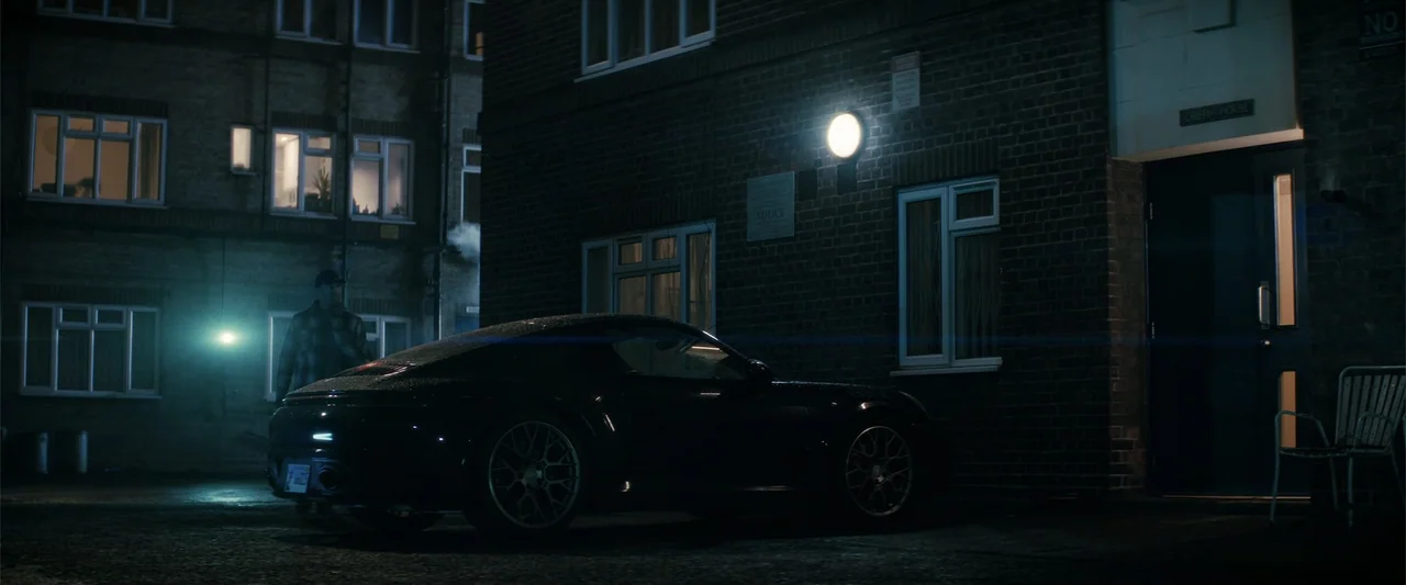 A Working Man (2025) movie still: wide — A dark brick apartment building stands at night with a parked car nearby. A solitary person stands in the…