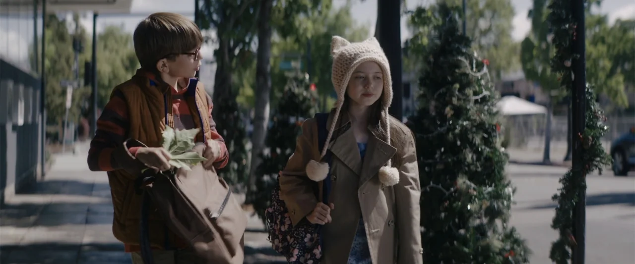 A Christmas Mystery (2022) movie still: medium — A sunny outdoor sidewalk is lined with trees and festive holiday greenery. A young boy and a young…