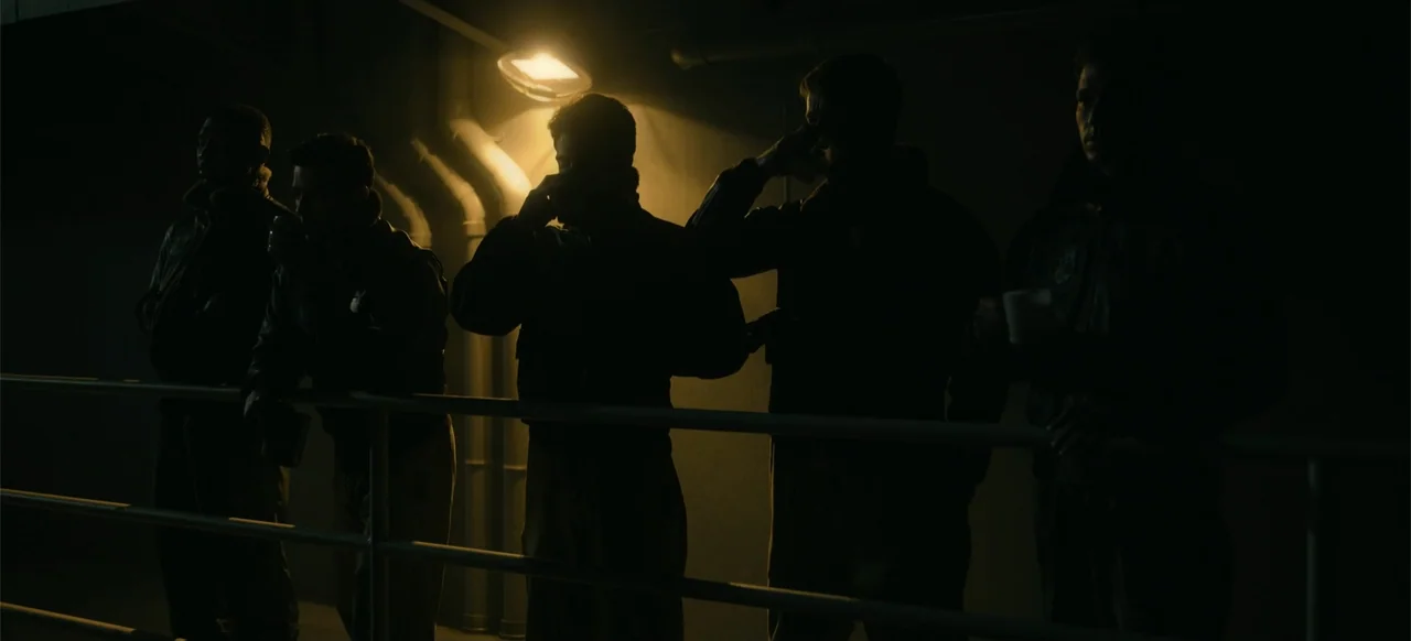 Devotion (2022) movie still: medium — Five pilots in flight gear stand in a dark, industrial corridor illuminated by overhead light. They are lined…