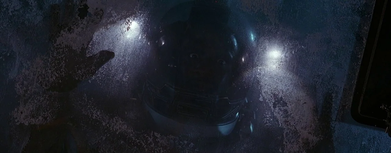Event Horizon (1997) movie still: closeup — A person in a space helmet looks through a frost-covered viewport inside a cockpit. The person presses a…