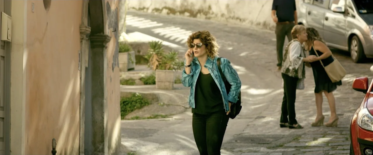 Taranta On the Road (2017) movie still: medium — A woman walks down a sunlit cobblestone street in a historic town. In the background two women…