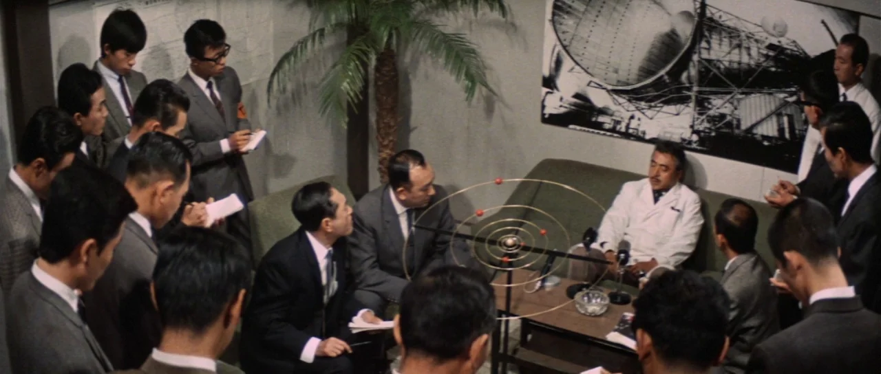 Invasion of Astro Monster (1965) movie still: wide — A crowded office space with a group of men sitting and standing around a table. A man in a…