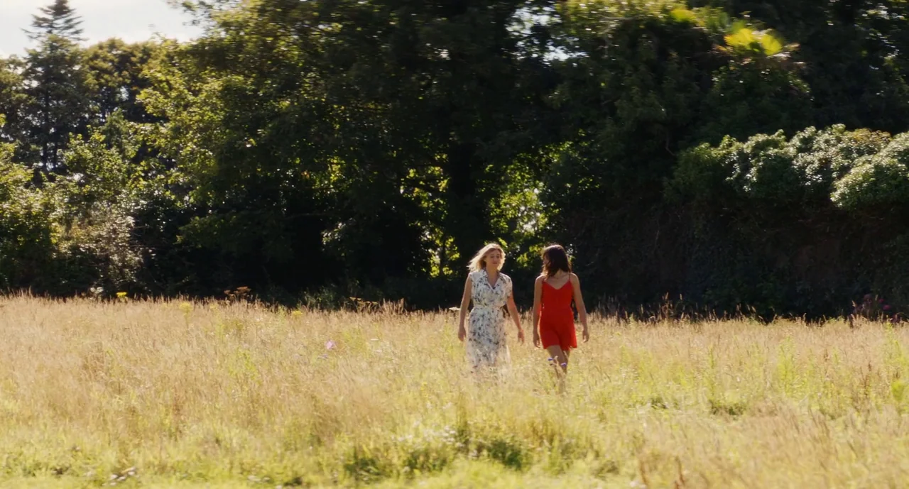 Anaïs in Love (2021) movie still: fullbody — A sunny meadow filled with tall golden grasses sits in front of a dense treeline. Two young women walk…