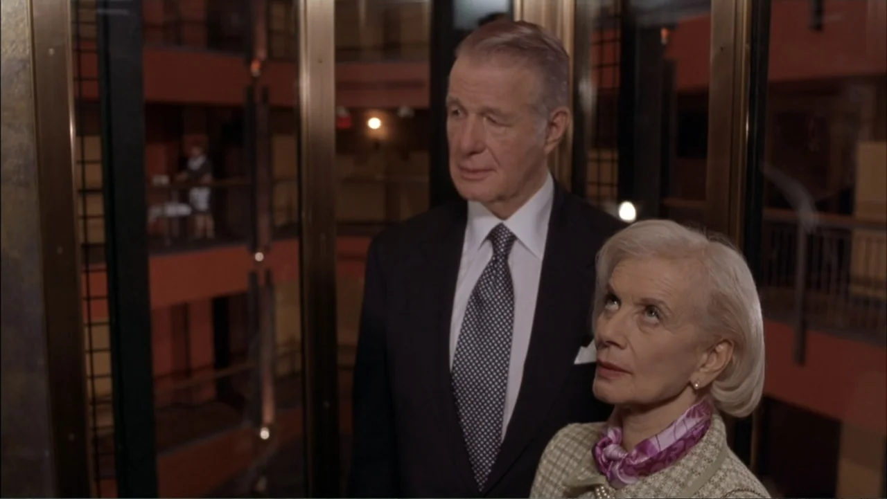 Two Weeks Notice (2002) movie still: medium — An older couple stands inside a glass elevator with orange building walls in view. The man wears a…