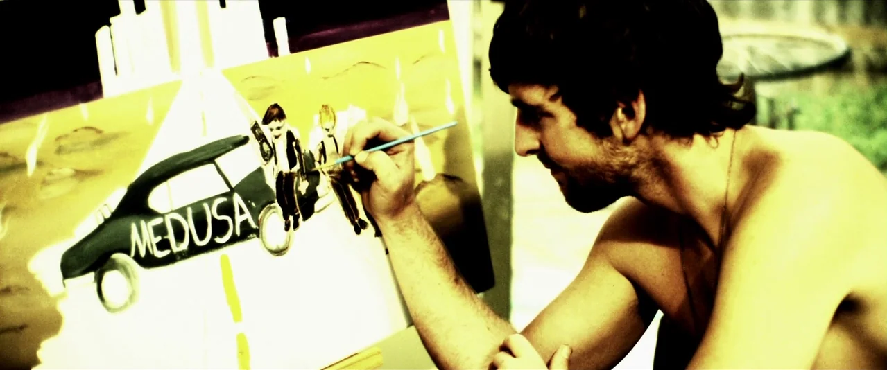 Bellflower (2011) movie still: medium — A young man stands beside an easel in a workspace with a yellow-tinted palette. He concentrates while…
