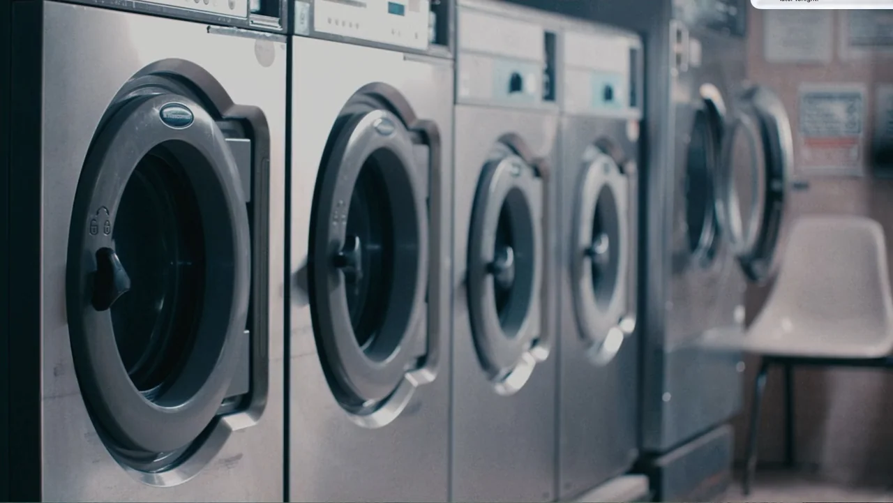 Terrifier 2 (2022) movie still: medium — A row of industrial stainless steel washing machines lines a brightly lit public space. A single white…