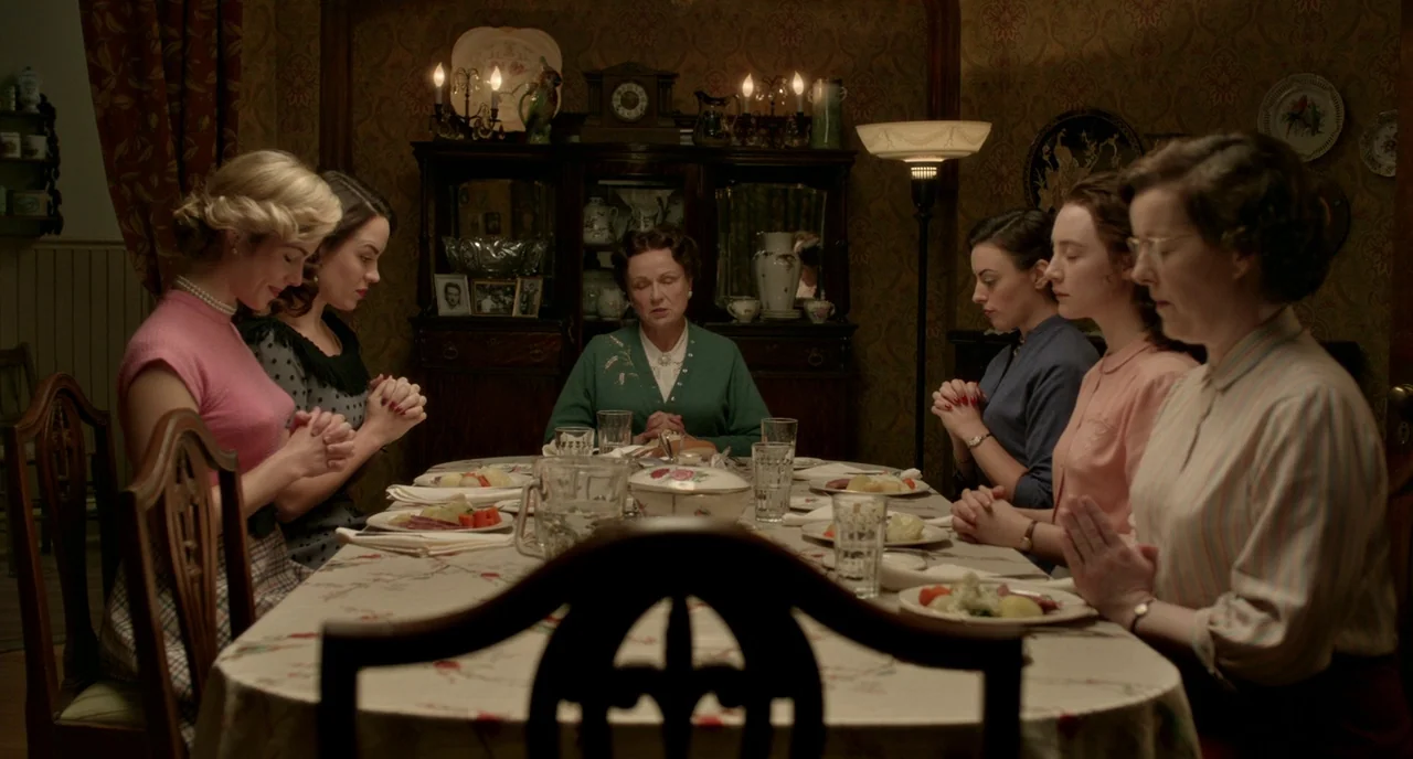Brooklyn (2015) movie still: medium — A 1950s dining room features patterned wallpaper and an ornate wooden cabinet holding dishes. Six women sit…