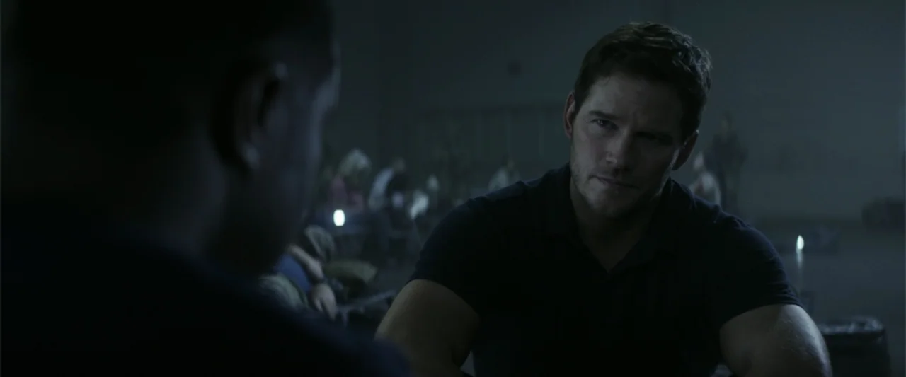 The Tomorrow War (2021) movie still: medium — A dark, expansive warehouse interior with rows of seats populated by blurred figures. A man sits in…